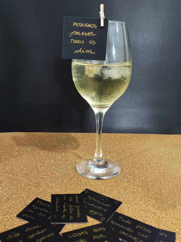 Copo de vinho branco com cubos de gelo e a mensagem "Pequenos passos todos os dias" escrita em um bilhete preto fixado no copo.