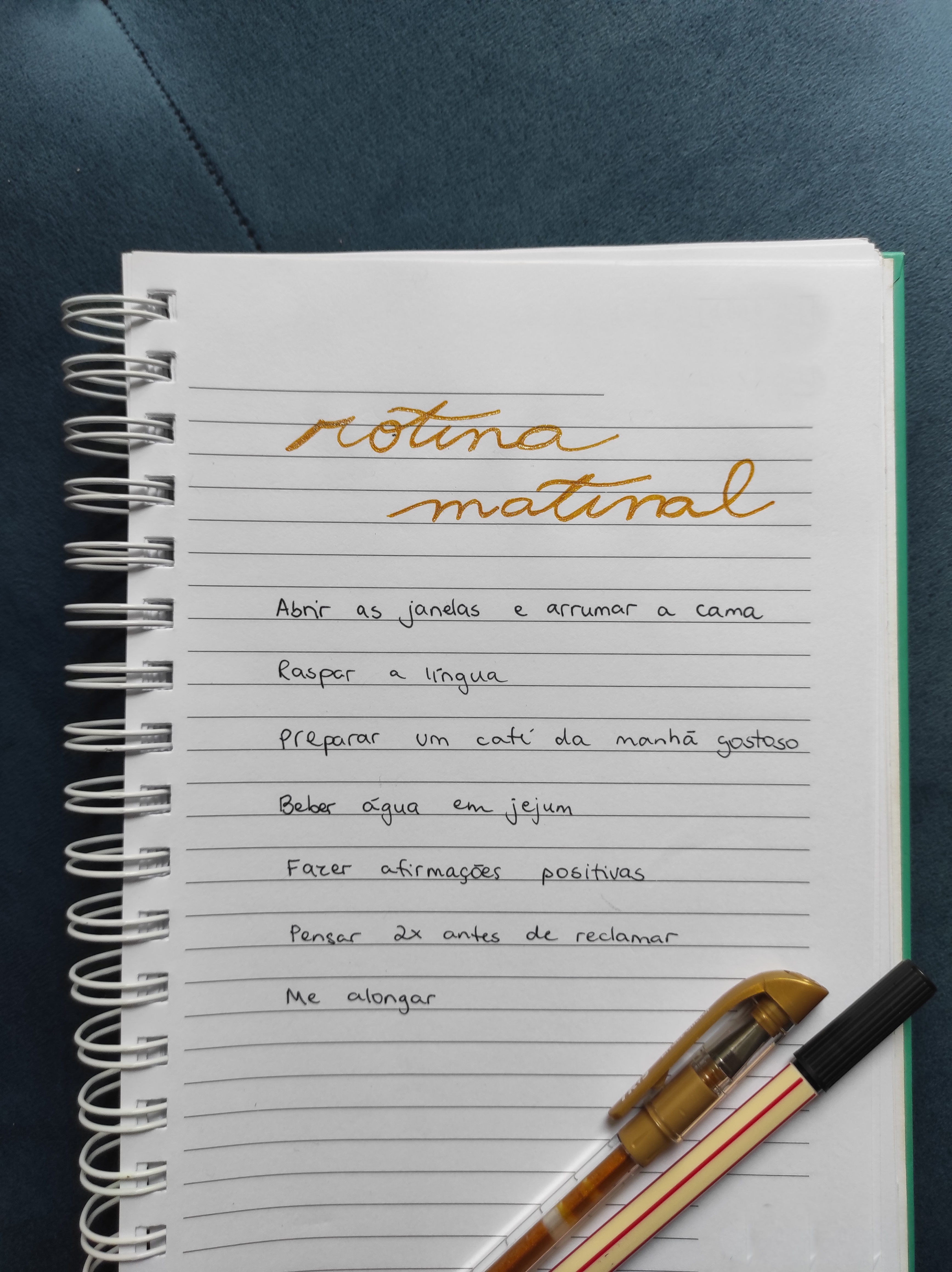 Caderno com uma lista escrita nas cores dourada e preta. O título é "rotina matinal" e os itens da lista são "Abrir as janelas e arrumar a cama, raspar a língua, preparar um café da manhã gostoso, beber água em jejum, fazer afirmações positivas, pensar 2x antes de reclamar, me alongar".