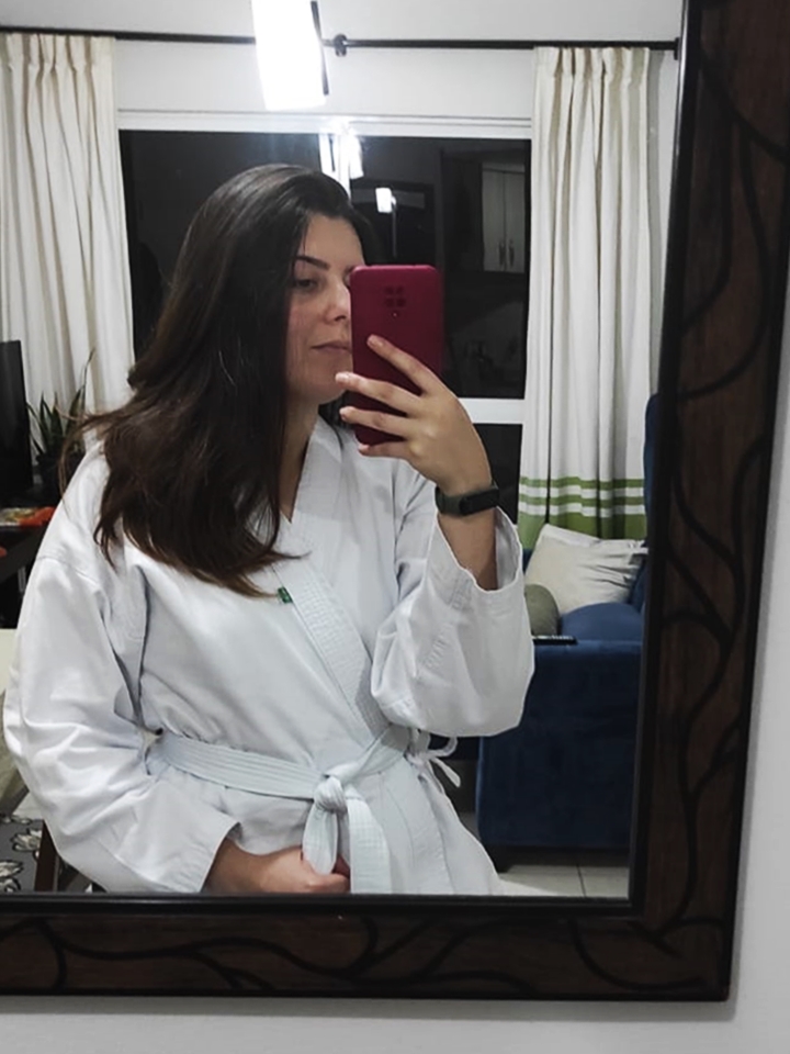 Mulher de cabelos castanhos com um kimono branco de artes marciais, tirando foto contra o espelho.