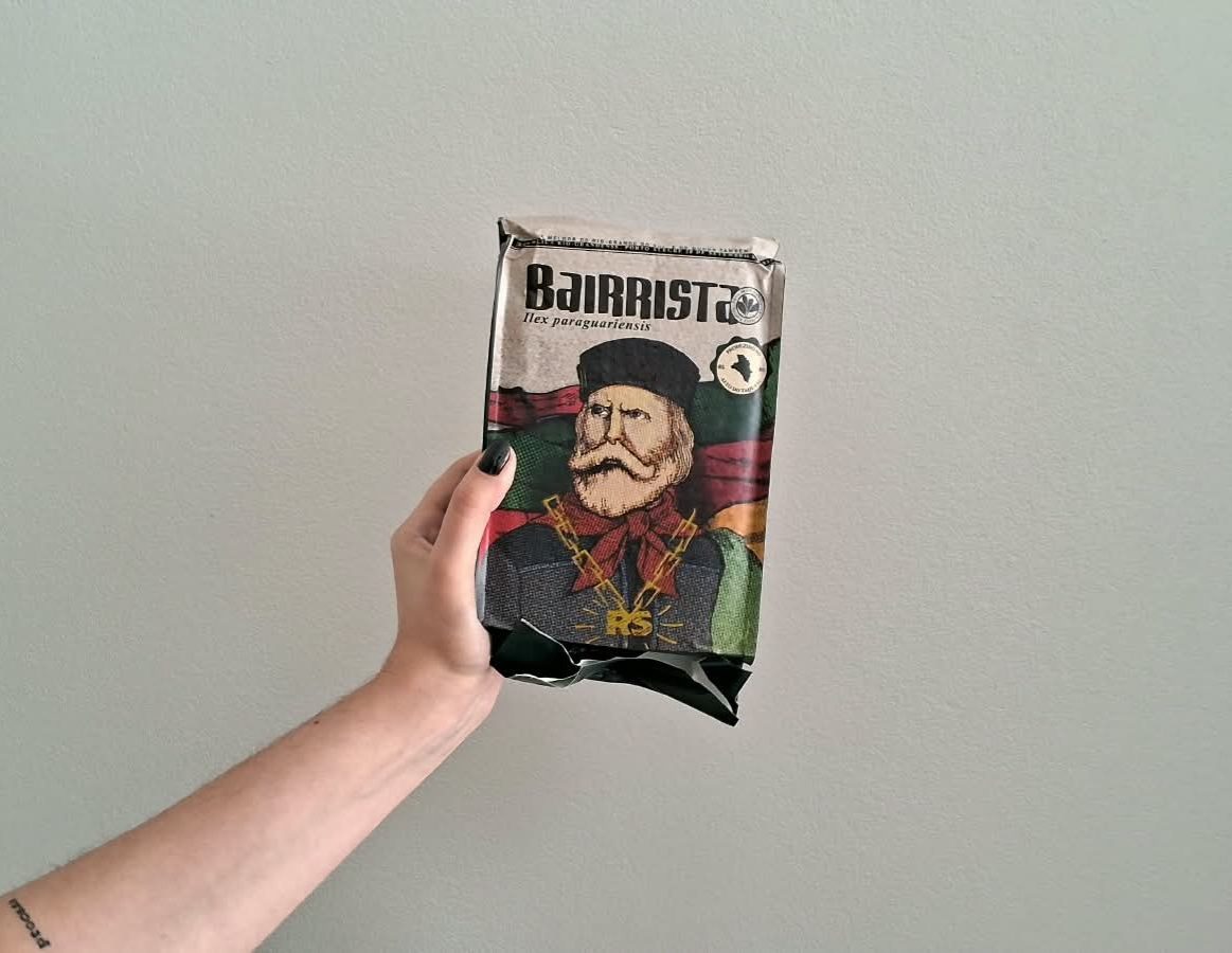 Uma mão segurando um pacote de erva da marca bairrista. O design da embalagem tem a ilustração de um homem de barba e bigode brancos, vestido com trajes típicos gaúchos e ao fundo as cores da bandeira do Rio Grande do Sul.